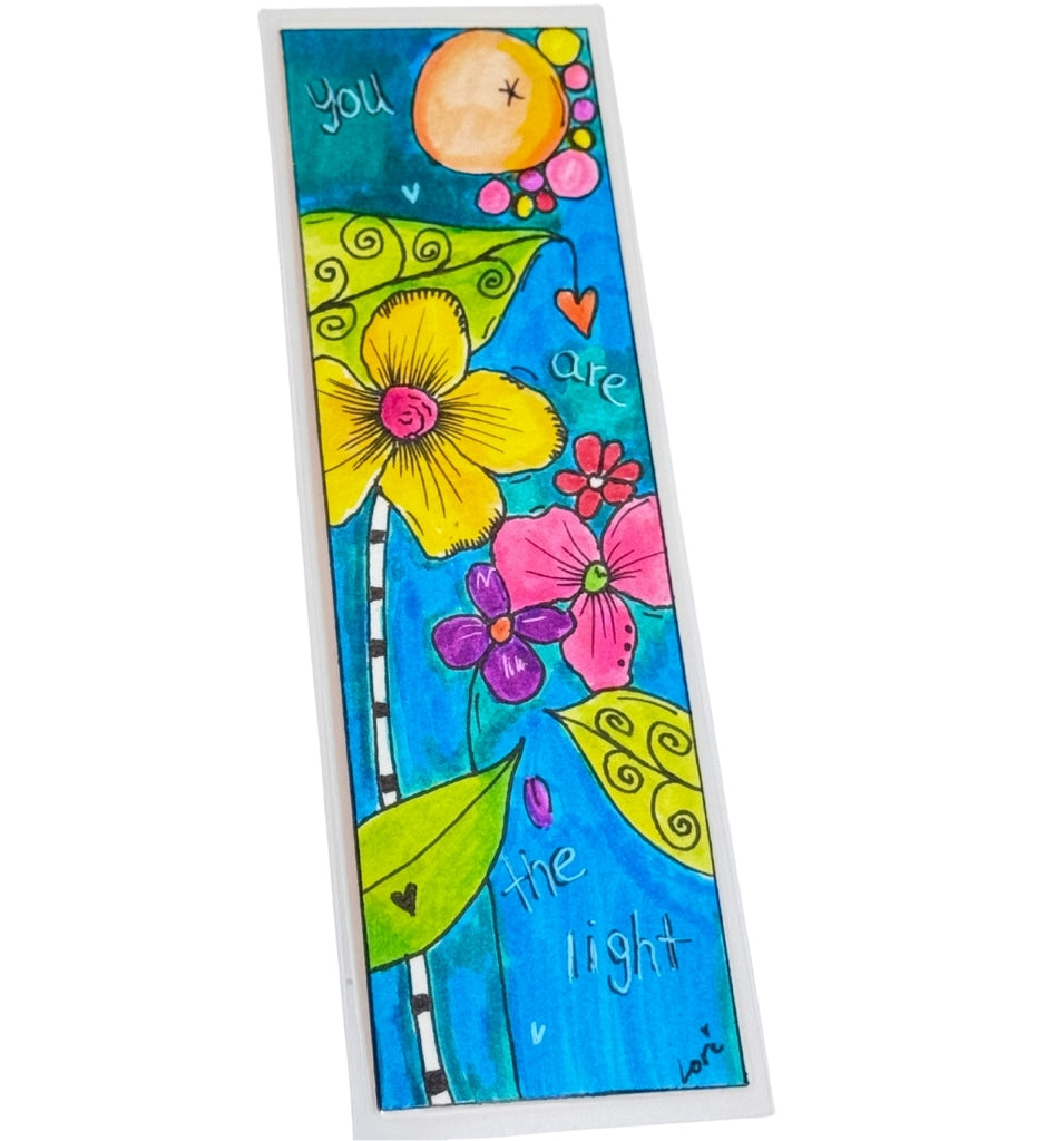 A colorful bookmark that is laminated featuring a whimsical floral design with yellow, pink, and purple flowers, green leaves, and the handwritten text 'you are the light' on a bright blue background.