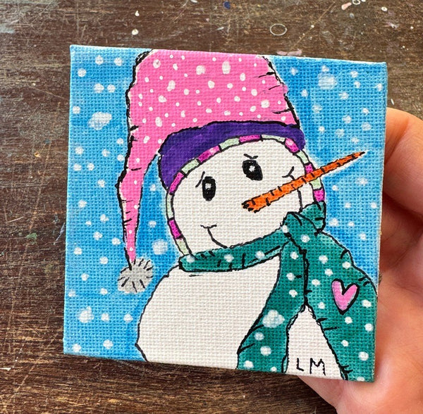 A small square canvas painting of a snowman wearing a pink hat and green scarf, held in a hand over a wooden surface.