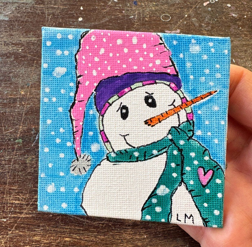 A small square canvas painting of a snowman wearing a pink hat and green scarf, held in a hand over a wooden surface.