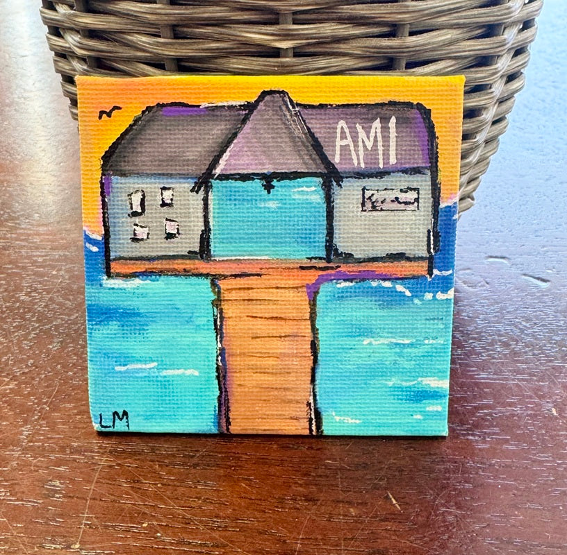 Small painting of building on a pier with 'AMI' with a sunset background placed on a wooden surface.