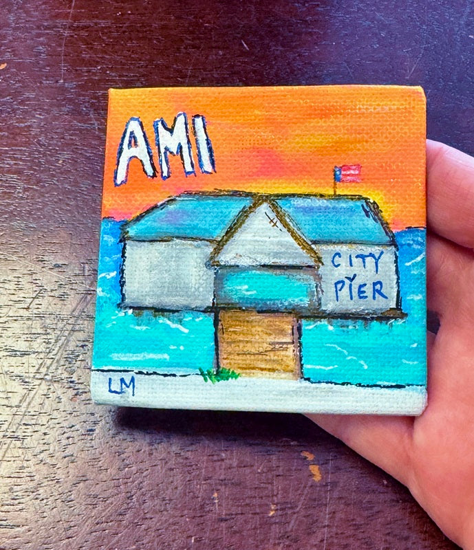 Small painted artwork of a building with 'AMI' and 'City Pier' text, held by a hand on a wooden surface.