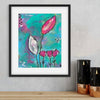 Framed abstract art print with pink flowers on a teal background, displayed on a wall above a wooden surface.