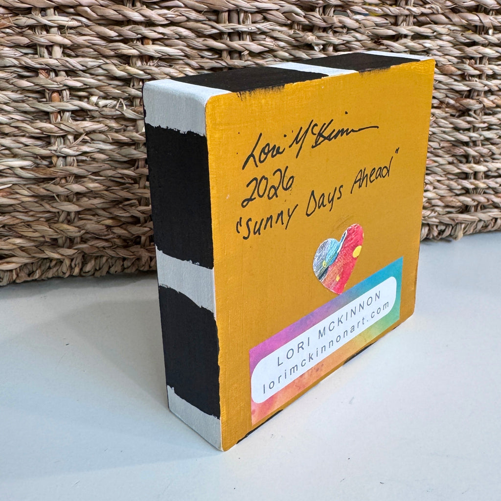 a wood block on a table in front of a basket. The block is painted and it has the artist name and date on it