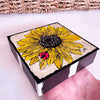 a wood block held by a hand with a sunflower painting on the front. There is part of a basket in the background
