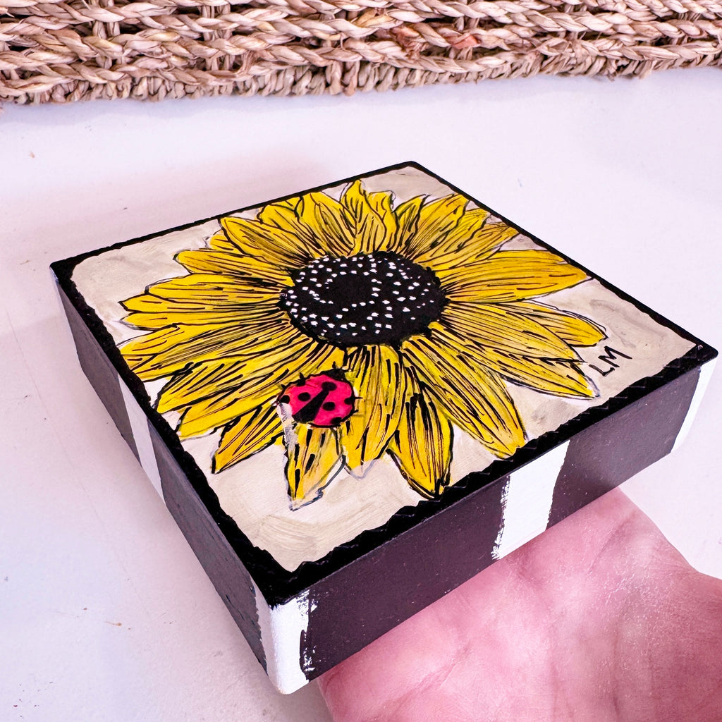 a wood block held by a hand with a sunflower painting on the front. There is part of a basket in the background
