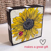 a wood block with a sunflower painting standing on a white table with a basket in the background. The sides of the block are painted black and white