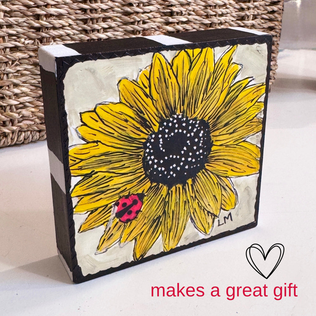 a wood block with a sunflower painting standing on a white table with a basket in the background. The sides of the block are painted black and white
