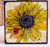 a wood block with a sunflower painting standing on a white table with a basket in the background