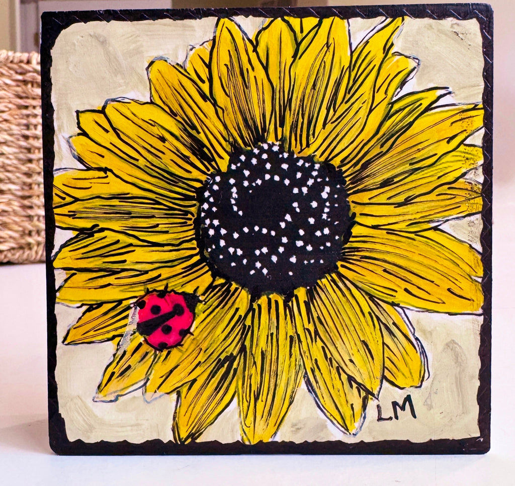a wood block with a sunflower painting standing on a white table with a basket in the background