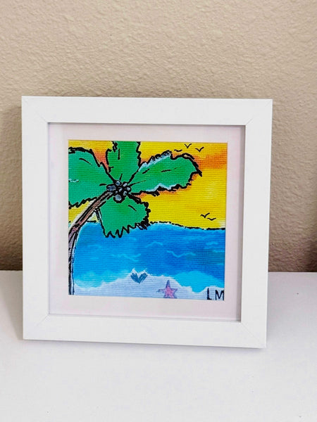 A framed print of a beach scene with a palm tree, blue ocean, yellow sky, and small birds, displayed on a white surface against a beige wall.