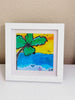 A framed print of a beach scene with a palm tree, blue ocean, yellow sky, and small birds, displayed on a white surface against a beige wall.