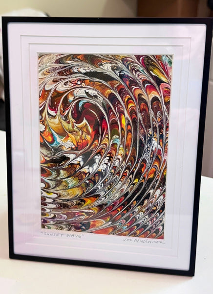 A framed art print featuring a swirling marbled pattern in many colors, displayed on a light surface with a black frame and white mat. Artist signature on the bottom