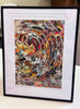 A framed art print featuring a swirling marbled pattern in many colors, displayed on a light surface with a black frame and white mat. Artist signature on the bottom