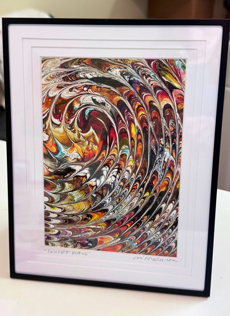 A framed art print featuring a swirling marbled pattern in many colors, displayed on a light surface with a black frame and white mat. Artist signature on the bottom