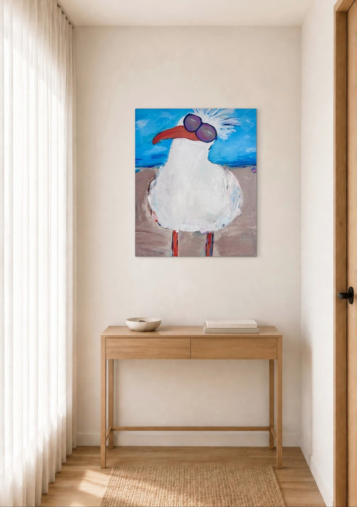 A canvas art print featuring a whimsical white seagull wearing purple sunglasses, hanging on a beige wall above a light wood console table in a bright, modern room.