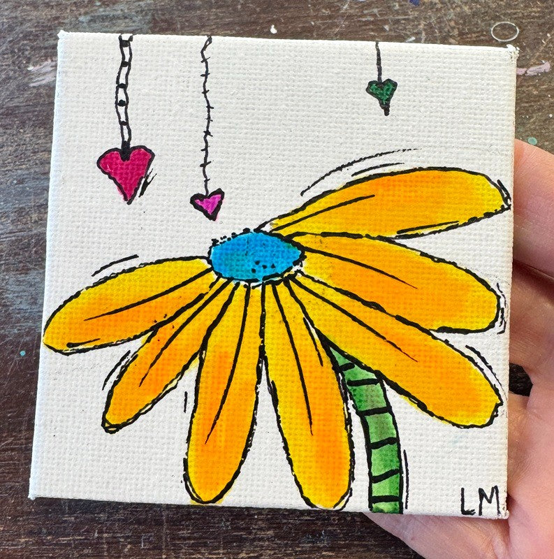 A small square canvas painting of a daisy with yellow-orange petals, a blue center, and a green stem, held by a hand over a dark wooden surface. Three hanging hearts appear above the flower.